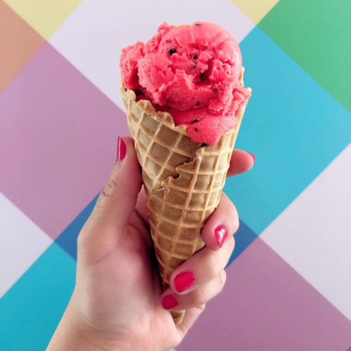 A cone of ice cream being held by a hand in front of a brightly coloured background
